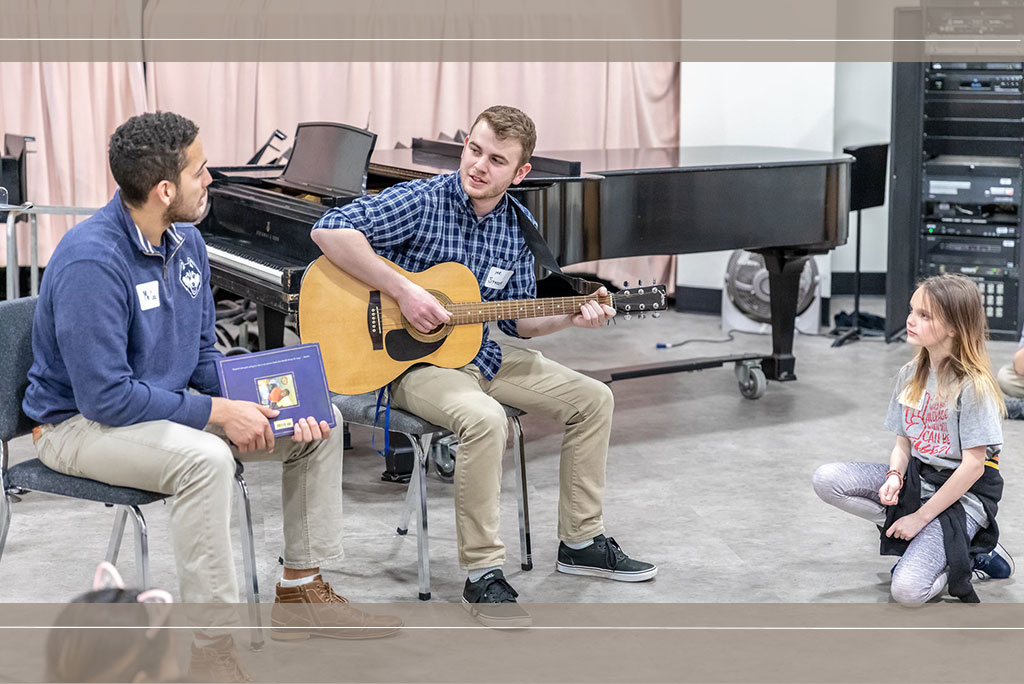 Two Neag School students with young local student in music room.